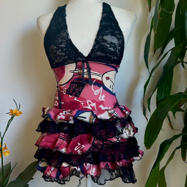 A sleeveless ruffled top displayed on a white mannequin torso, featuring a black lace halter-style upper section and a tiered skirt made from pink, red, white, and black patterned fabric with lace trim, set against a light wall with green leaves at the sides.