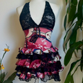 A sleeveless ruffled top displayed on a white mannequin torso, featuring a black lace halter-style upper section and a tiered skirt made from pink, red, white, and black patterned fabric with lace trim, set against a light wall with green leaves at the sides.
